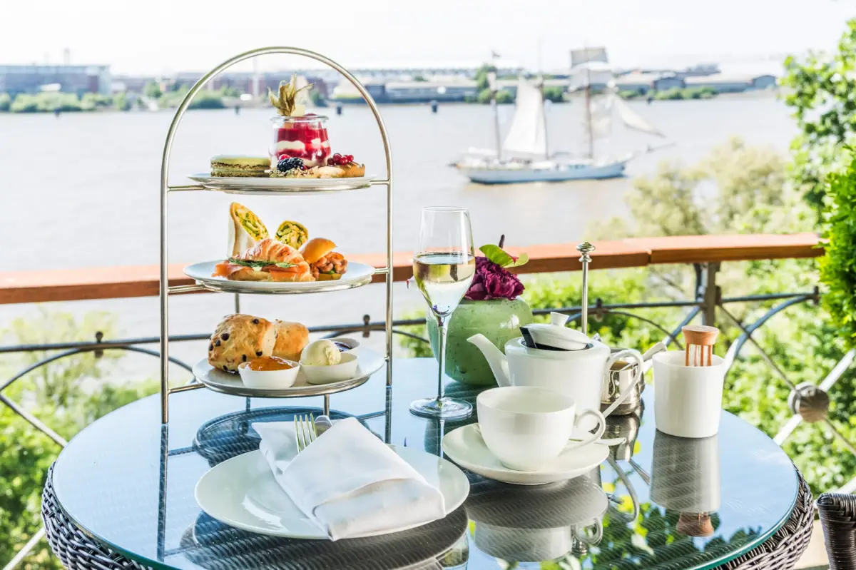 Afternoon Tea Lindenterrasse Englische Teezeit mit herzhaften und süßen Kleinigkeiten serviert auf einem Tisch auf der Lindenterrasse des Hotels Louis C. Jacob mit Blick auf die Elbe und ein Segelboot.
