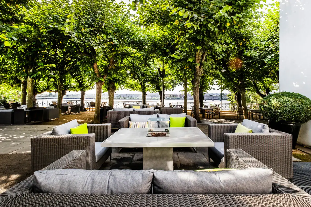 Lindenterrasse Tische und Sofas stehen unter freiem Himmel auf der Lindenterrasse mit grünen Linden im Hintergrund.