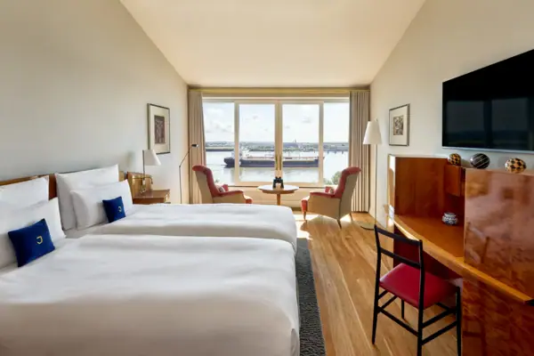 Deluxe Doppelzimmer Elbblick Ein Hotelzimmer mit einem Bett mit weißen Bettlaken und einem großen Fenster mit Blick auf die Elbe.