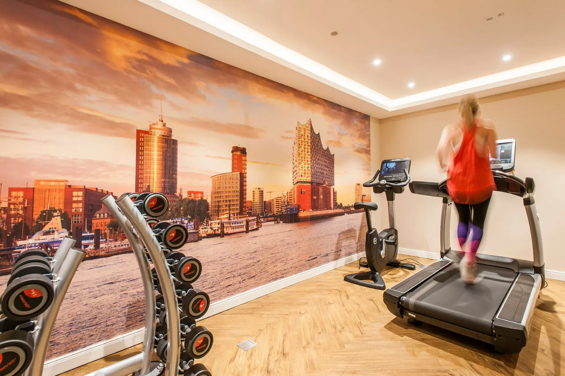 Fitness Eine Frau läuft auf einem Laufband im Fitness-Bereich des Louis C. Jacob in einem Raum mit einem großen Wandgemälde.