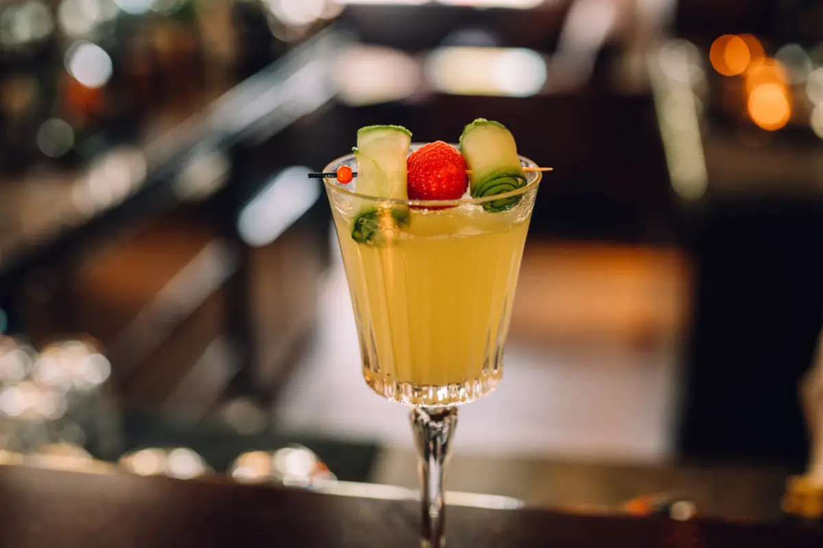 Drink Jacobs Bar Ein Cocktailglas mit einer gelben Flüssigkeit des Cocktails Mrs. Blankenese mit einer Garnitur von Gurke und Himbeere.