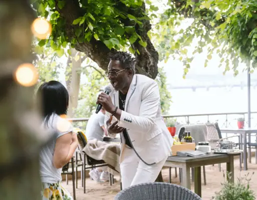 Ein Mann in einem weißen Anzug singt in ein Mikrofon unter einem Baum.