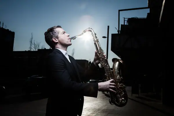 Toddi Reed Ein Mann in einem schwarzen Anzug spielt Saxophon und hinter ihm scheint die Sonne.