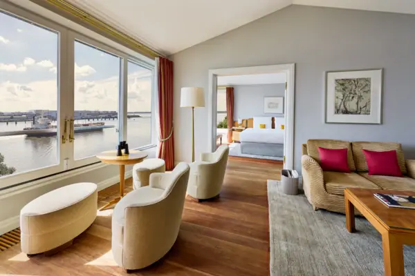 Suite Elbblick Ein großes Hotelzimmer mit großen Fenster und tollem Blick auf die Elbe sowie mit einem gemütlichen Wohnbereich im Vordergrund und einem separaten Schlafzimmer im Hintergrund.