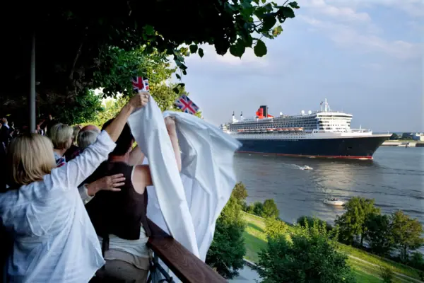 Verabschiedung der Queen Mary 2 auf der Lindenterrasse Eine Gruppe von Menschen hält Fahnen und winkt vor einem großen Schiff.
