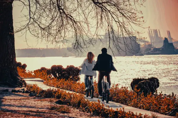 Ein Mann und eine Frau fahren Fahrrad im Herbst auf einem Radweg direkt an der Elbe mit Blick auf den Hamburger Containerhafen.