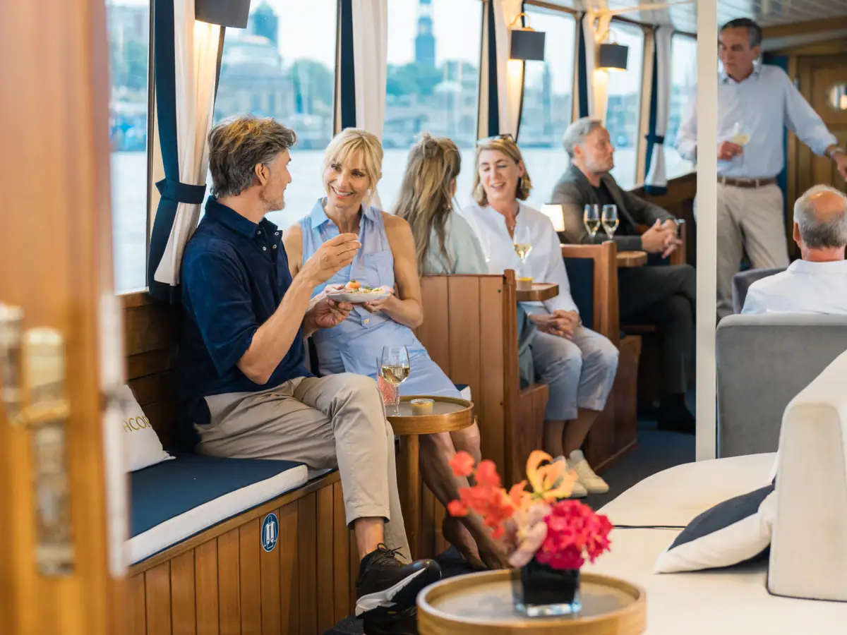 Mehrere Personen sitzen Unterdecks in der Barkasse MS Jacob, trinken Sekt und genießen Snacks.