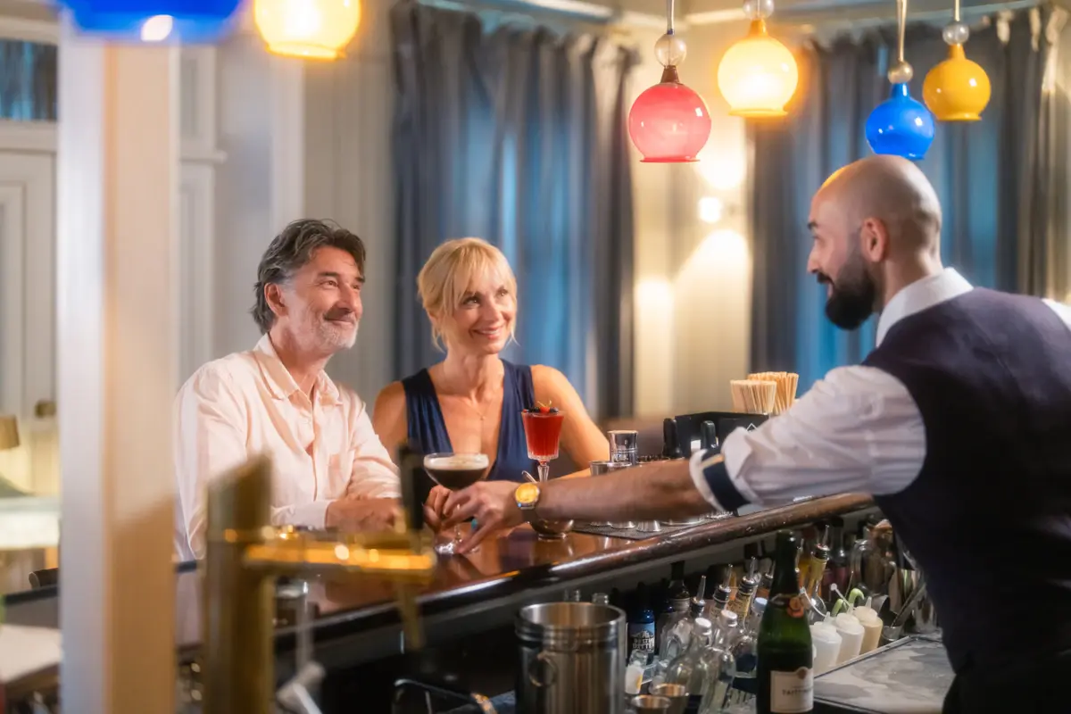 Jacobs Bar und Wohnhalle Zwei Menschen sitzen an der Jacobs Bar und der Barkeeper stellt ihnen Cocktails auf die Theke.