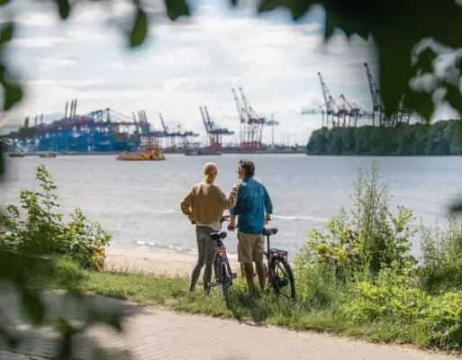 Ein Mann und eine Frau stehen mit Fahrrädern am Elbstranmd und schauen über die Elbe auf den Containerhafen Hamburg.