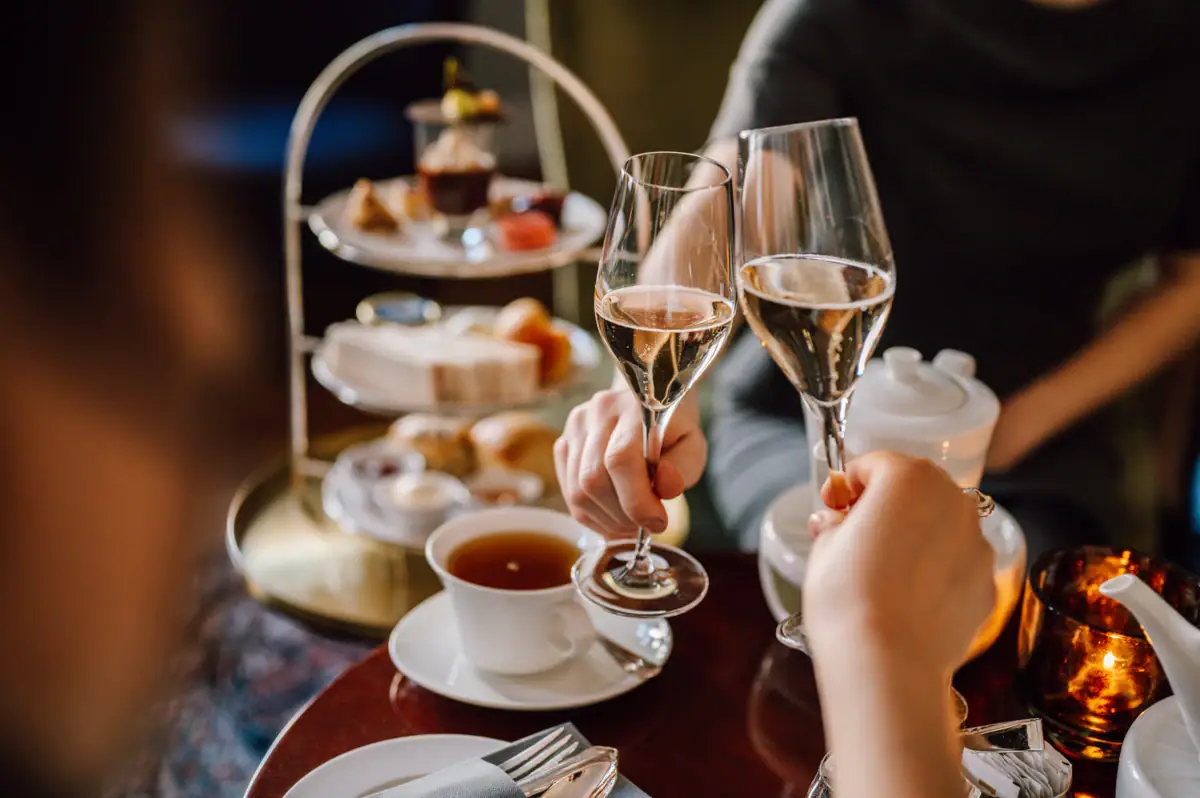 Afternoon Tea Hamburg Teezeit im Hotel Louis C. Jacob. Zwei personen stoßen mit Champagner an und im Hintergrund sieht man eine Etagere mit herzhaften und süßen Köstlichkeiten.