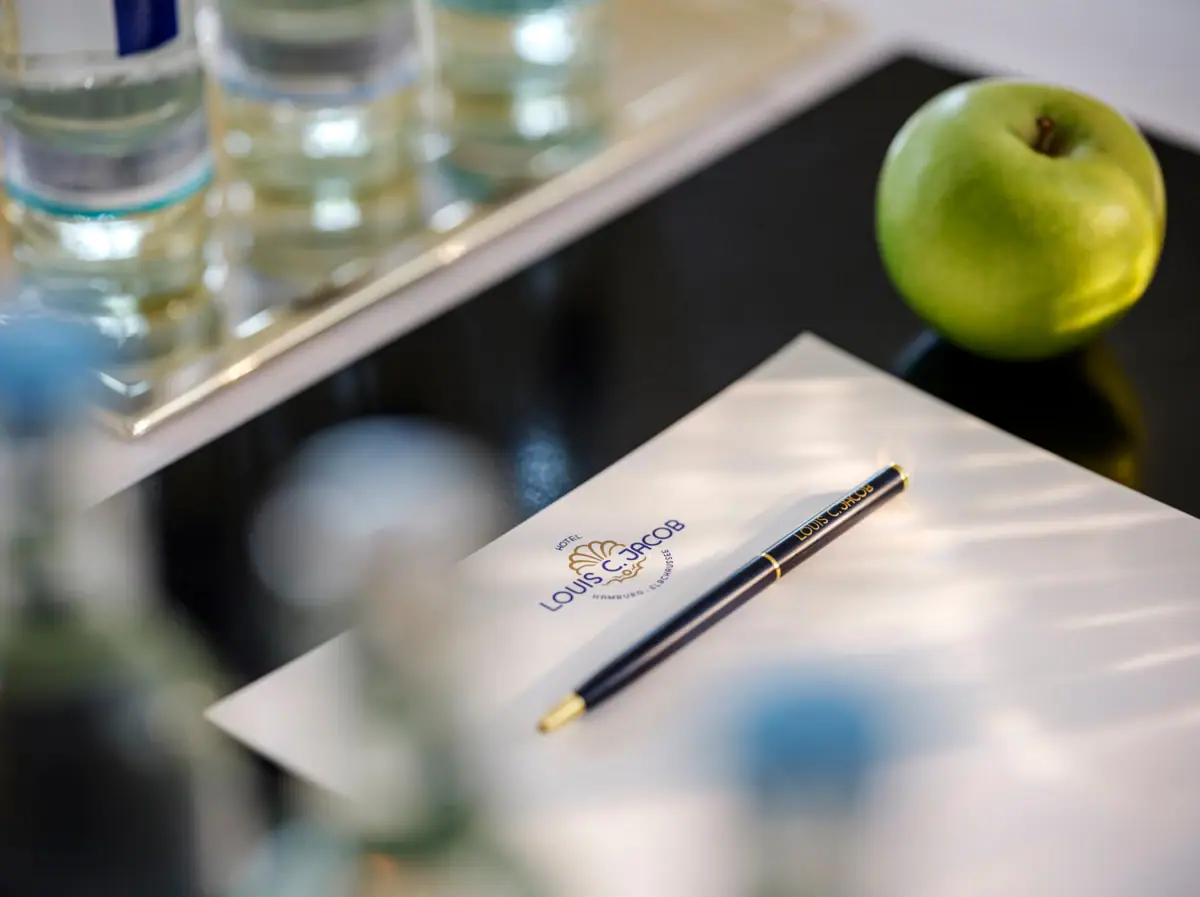 Platz Setup bei einer Tagung Auf einem Tisch liegt ein Kugelschreiber und Schreibblock des Hotels Louis C. Jacob neben einem Apfel.