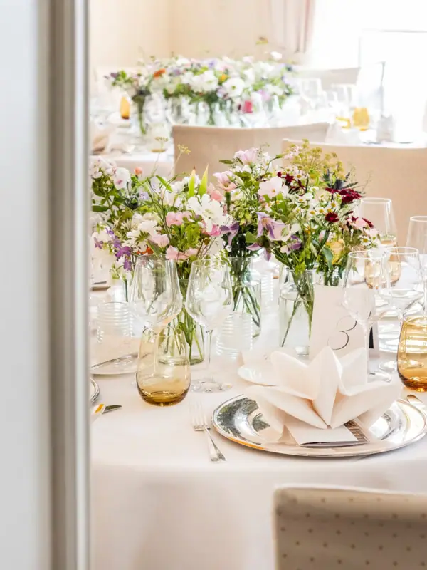 Ein Tisch mit Blumenarrangement als Mittelpunkt, umgeben von Tischgeschirr und Tischdecke.